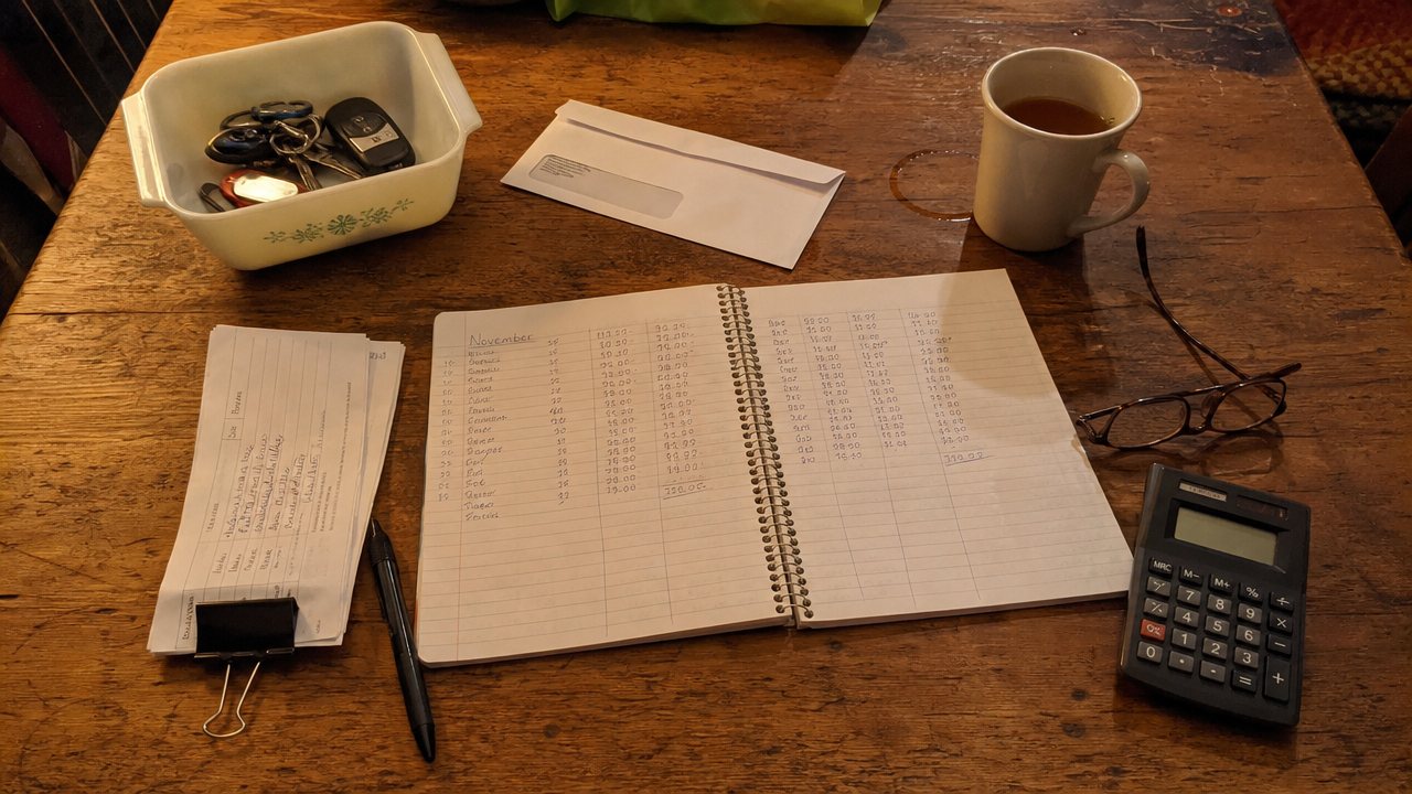 Kitchen table with a handwritten sales ledger, calculator, and booth sales slips for a monthly budget check.