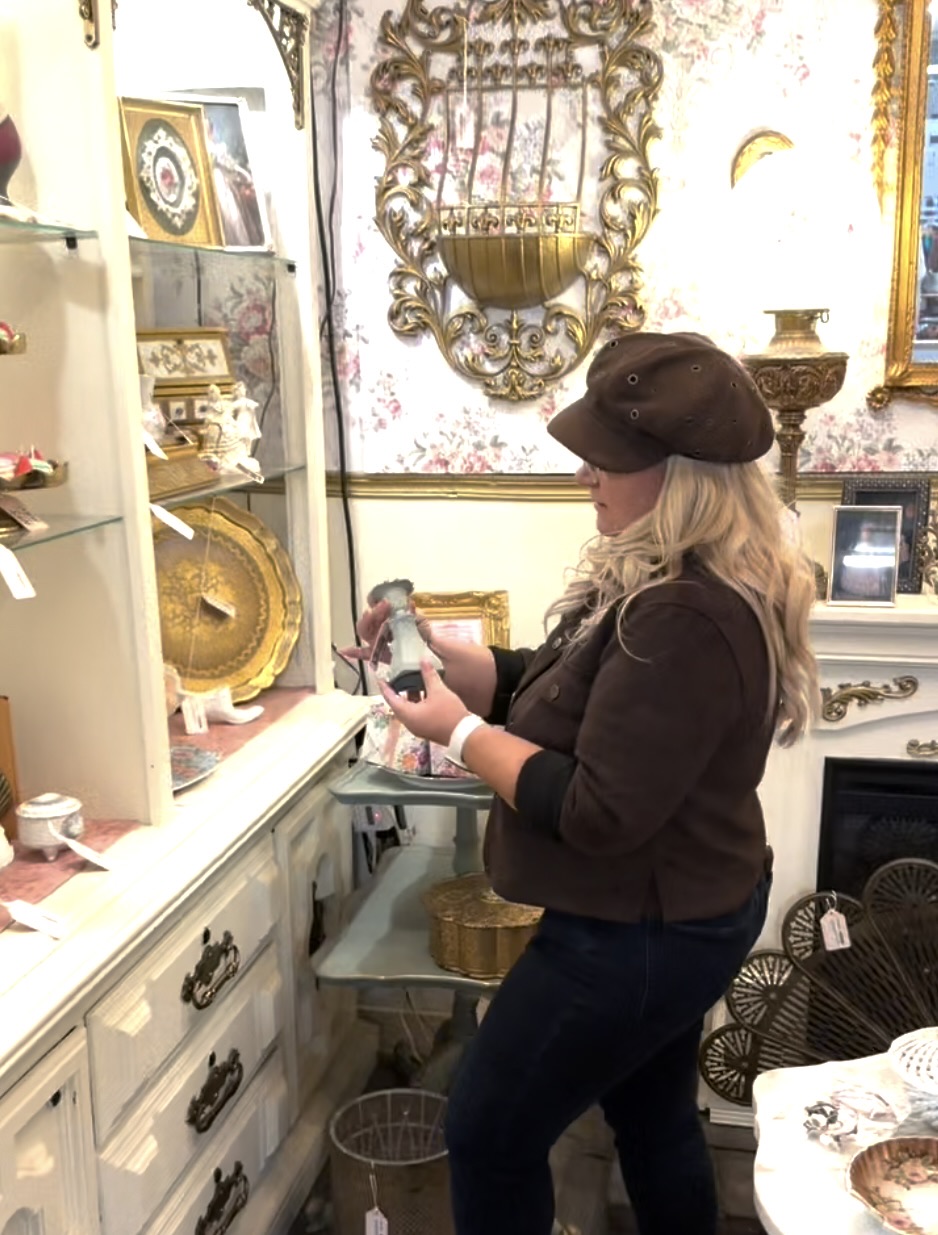 A shopper in a brown newsboy cap examines a porcelain figurine in an antique mall booth with floral wallpaper, gold-framed mirrors and vintage decor
